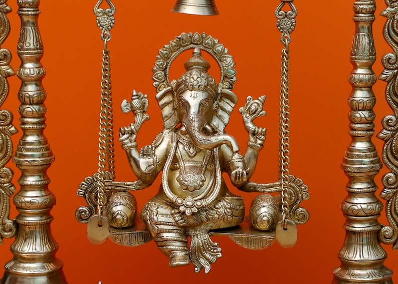 May include: A gold-coloured metal statue of Ganesha, the Hindu god of wisdom and prosperity, sitting on a swing. The statue is intricately detailed and has a traditional design.