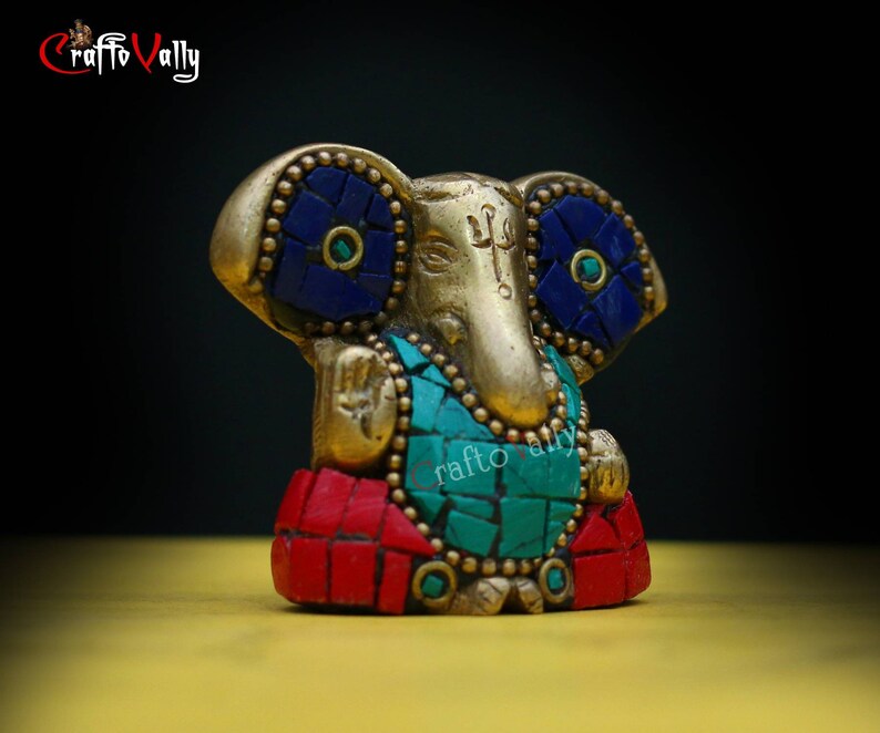Puede incluir: Una estatua dorada de Ganesha con detalles en mosaico rojo, azul y verde. La estatua est&aacute; sentada sobre una superficie amarilla.