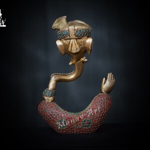 May include: A gold-colored brass statue of Ganesha, the Hindu god of wisdom and prosperity. The statue is decorated with red and green gemstones. The statue is in a sitting position with one hand raised in a blessing gesture.