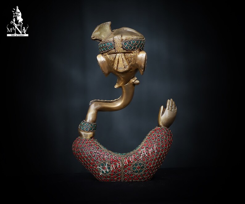 May include: A gold-colored brass statue of Ganesha, the Hindu god of wisdom and prosperity. The statue is decorated with red and green gemstones. The statue is in a sitting position with one hand raised in a blessing gesture.