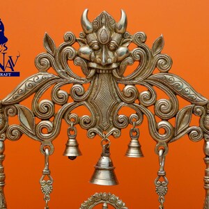 May include: A silver-coloured metal decorative stand with intricate floral designs and a central motif of a demon face. The stand has several hanging bells.