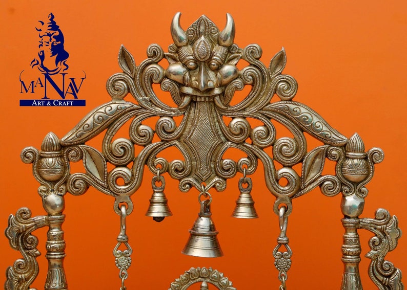 May include: A silver-coloured metal decorative stand with intricate floral designs and a central motif of a demon face. The stand has several hanging bells.