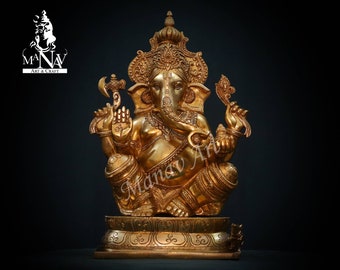 May include: A gold-colored metal statue of Ganesha, the Hindu god of wisdom and prosperity. The statue is sitting on a base with intricate details. The statue is adorned with ornate jewelry and has a trunk that is curled to the left.