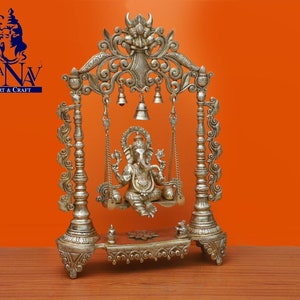May include: Silver-toned metal statue of Ganesha, the Hindu god of wisdom and prosperity, sitting on a swing. The swing is decorated with intricate carvings and bells. The statue is on a base with a floral design.
