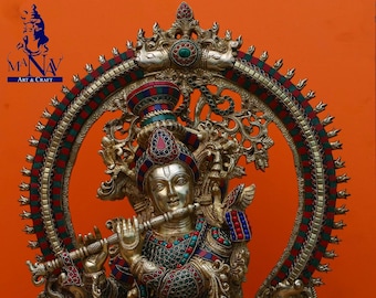 Op de afbeelding: Een gouden standbeeld van Krishna die fluit speelt, versierd met kleurrijke edelstenen en ingewikkelde details. Het beeld wordt omringd door een cirkelvormige halo met een decoratief ontwerp.