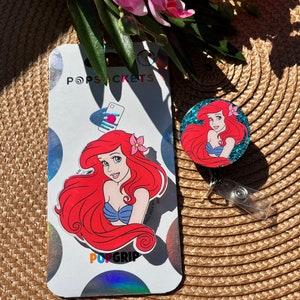 May include: A red and blue glitter phone grip with a picture of Ariel from The Little Mermaid. The grip is attached to a clear plastic badge holder with a metal clip. The packaging says "PopSockets" and "PopGrip".