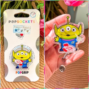 May include: A clear plastic phone grip with a green alien character from the movie Toy Story. The alien is wearing a blue jumpsuit and holding a red and white pizza planet logo. The phone grip is attached to a retractable badge reel.