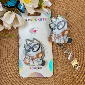 May include: A white and brown cartoon dog with glasses and a brown scarf is featured on a phone grip and a retractable badge holder. The phone grip is on a white card with the text "POPGRIP" and "OPSOCKETS".