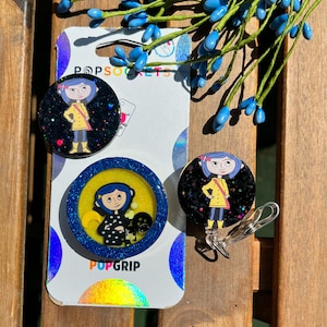 May include: Two round phone grips featuring a cartoon character from the movie "Coraline". The grips are black with glitter and feature a yellow background with glitter. The text "PopGrip" is printed on the bottom of one of the grips. The text "PopSockets" is printed on the top of the other grip.