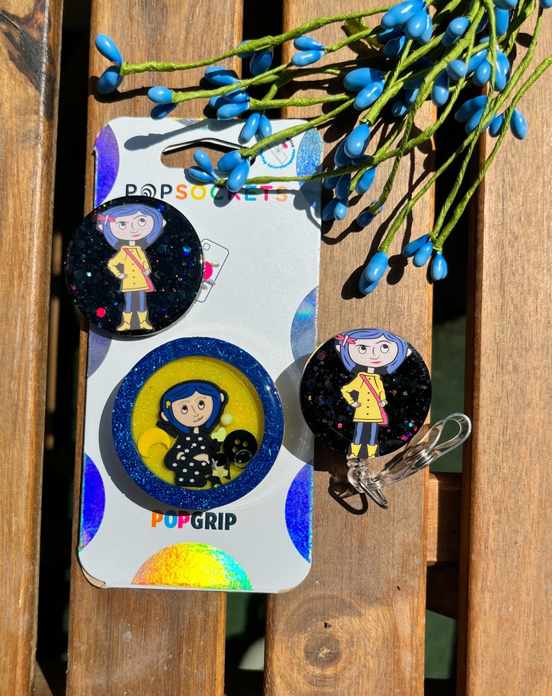 May include: Two round phone grips featuring a cartoon character from the movie "Coraline". The grips are black with glitter and feature a yellow background with glitter. The text "PopGrip" is printed on the bottom of one of the grips. The text "PopSockets" is printed on the top of the other grip.