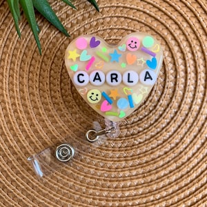 May include: A clear heart-shaped resin keychain with a gold glitter background and colorful embellishments. The name "CARLA" is spelled out in white letters.