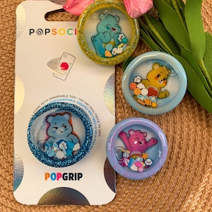 May include: Three Care Bear character phone grips with glitter and colorful accents. The grips are blue, yellow, and light blue. The text "POPSOCI" and "POPGRIP" are on the packaging.