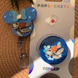 May include: A blue and white glitter phone grip with a cartoon turtle design. The grip is attached to a blue and white glitter phone charm with a cartoon turtle design. The charm is attached to a retractable phone strap. The packaging says "PopSockets" and "PopGrip".