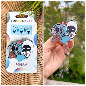 May include: A blue and white heart-shaped PopGrip with a cartoon image of two robots holding hands. The PopGrip is attached to a clear plastic badge holder with a retractable cord. The packaging includes the text "PopSockets", "Roomocrafts by Carla", and "PopGrip".