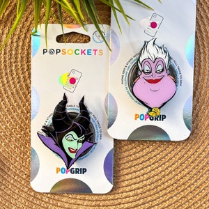 May include: Two phone grip and stand accessories featuring Disney characters. The first features Ursula from The Little Mermaid with a purple and pink design. The second features Maleficent from Sleeping Beauty with a black, green, and purple design. Both have the text "PopGrip" on the bottom.