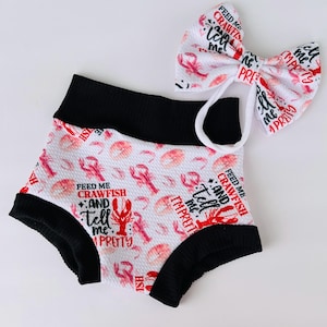 May include: White baby shorts and matching bow with a lobster print and the text "Feed Me Crawfish and Tell Me I'm Pretty". The shorts have black trim.
