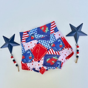Pode incluir: Calções de cintura alta com um design de patchwork com bolsos com aspeto de ganga, riscas vermelhas e padrões de morangos. Os calções são decorados com detalhes em azul, vermelho e branco. Enfeites decorativos de estrelas em ambos os lados.