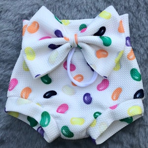 May include: White baby bloomers with a large jelly bean print and a matching jelly bean bow. The bow is tied with a white elastic.