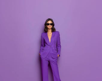 May include: A woman wearing a vibrant purple suit with a tailored blazer and matching wide-leg pants. The suit features a plunging neckline and is paired with black sunglasses. The background is a solid purple.
