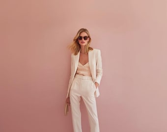 May include: A cream-colored pantsuit with a matching blazer and trousers. The blazer has a classic cut, and the trousers are straight-legged. A light peach-colored camisole is worn underneath. The person is wearing sunglasses and holding a small clutch. The background is a solid pink.