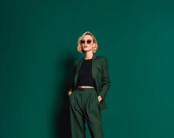 May include: A person wearing a dark green suit with a black top and sunglasses. The suit includes a blazer and matching trousers. The background is a solid dark green.