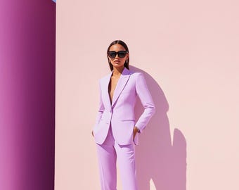 May include: A light purple suit with a tailored blazer and matching pants. The suit is worn with sunglasses. The background is a light pink wall with a purple wall on the left side.