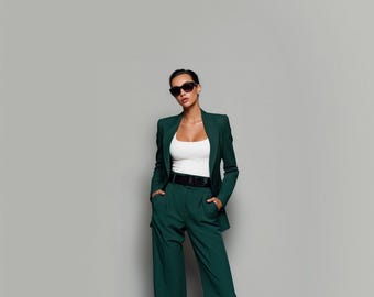 May include: A dark green pantsuit with a matching blazer and wide-leg trousers. The blazer is worn over a white top and a black belt. The person is wearing black sunglasses.
