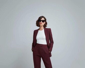 May include: A burgundy suit, consisting of a blazer and matching trousers, paired with a white top and black sunglasses. The blazer has a tailored fit, and the trousers have a straight-leg design. The outfit is set against a plain gray background.
