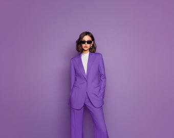 May include: A person wearing a purple suit with a white top and black sunglasses. The suit includes a blazer and wide-leg pants. The background is a solid purple.
