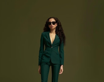 May include: A dark green suit with a single-button blazer and matching trousers. The blazer has a notched lapel and a tailored fit. The person in the image is wearing black sunglasses. The background is a solid, olive green color.