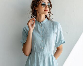 May include: A light blue linen dress with a button-up front and short sleeves. The dress has a relaxed fit and a slightly flared skirt. The model is wearing round sunglasses.