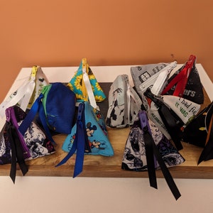 May include: A collection of 12 small triangular zippered pouches made of fabric. The pouches are in various colors and patterns, including black, white, blue, green, yellow, and red. Some of the pouches have a character from a popular cartoon printed on them. The pouches are arranged in a pyramid shape on a wooden surface.
