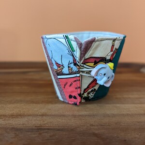 May include: A fabric cup cozy with a colorful comic book print featuring an elephant, a red and white striped tent, and the text "Three Cheers!" The cozy is teal green on the back side.