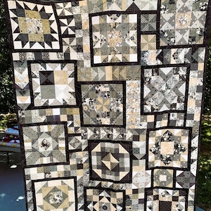 May include: A large, handmade quilt with a patchwork design. The quilt features various geometric patterns in shades of black, gray, and beige, with a dark brown border. The quilt is hanging outside, showcasing its intricate details and craftsmanship.
