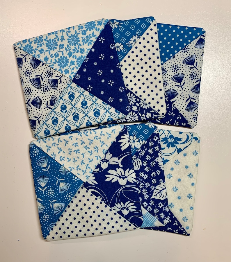 May include: Four blue and white fabric coasters with a patchwork design. Each coaster is made of different blue and white patterned fabrics sewn together in a square shape.