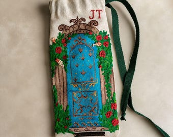 May include: A beige drawstring pouch featuring an embroidered design of a bright blue arched door surrounded by green vines and red flowers. The letters JT are stitched in red at the top. The pouch includes a long, dark green fabric strap.