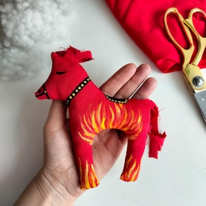 May include: A red fabric horse ornament featuring a painted yellow and orange flame pattern on its body. The horse has a black bridle and saddle detail with yellow dots, and a simple black line for an eye. The fabric edges are frayed.