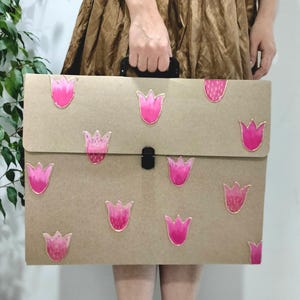 May include: A brown portfolio with a black handle and clasp, adorned with pink tulip designs outlined in gold. The portfolio is held by a person. The tulips have varying shades of pink and some have glitter accents.