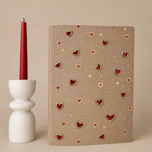 May include: A red candle stands in a white, sculptural candle holder next to a notebook. The notebook has a brown cover adorned with red and gold heart-shaped embellishments. The background is a neutral beige.
