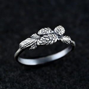 May include: A silver ring with a pine cone design. The ring is made of sterling silver and features two pine cones with needles.