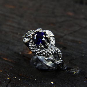 Może przedstawiać: Srebrny pierścionek z ciemnoniebieskim kamieniem szlachetnym osadzonym w wzorze pazura smoka. Pierścionek jest ozdobiony misternymi rzeźbami i fakturowanym wykończeniem.