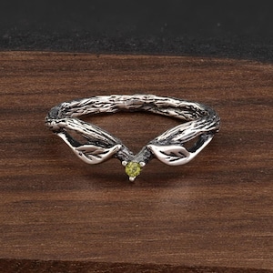 May include: A silver ring with a nature-inspired design. The ring has a textured band like tree bark, with leaf-like details either side of a small, round, light green gemstone. The ring is set against a dark wood grain background.