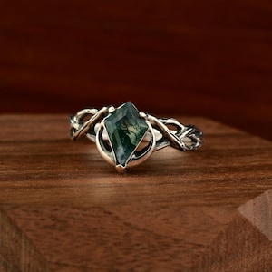 May include: A silver ring with a green moss agate gemstone in a diamond shape. The ring has a detailed, intertwined band design. The gemstone is approximately 1.3 cm in length.