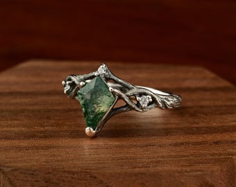 May include: A silver ring with a green moss agate gemstone. The ring has a nature-inspired design with a branch-like band and small, clear accent stones. The gemstone is a unique, angular shape. The ring is displayed on a wooden surface.