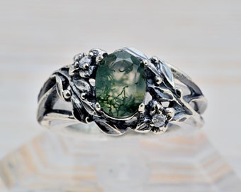 May include: A silver ring featuring a green moss agate stone set within a floral design. The ring is adorned with delicate silver leaves and flowers, accented by small clear stones.