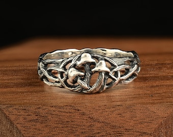 Puede incluir: Un anillo plateado con un diseño de setas. El anillo tiene una banda calada detallada con tres figuras de setas prominentes en el centro. El anillo está sobre una superficie de madera.