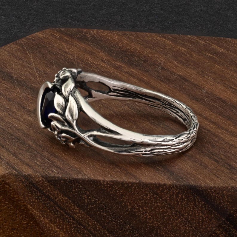 May include: A silver ring with a dark blue oval gemstone. The ring features a nature-inspired design with leaves and a textured band resembling a tree branch. The ring is set on a wooden surface.