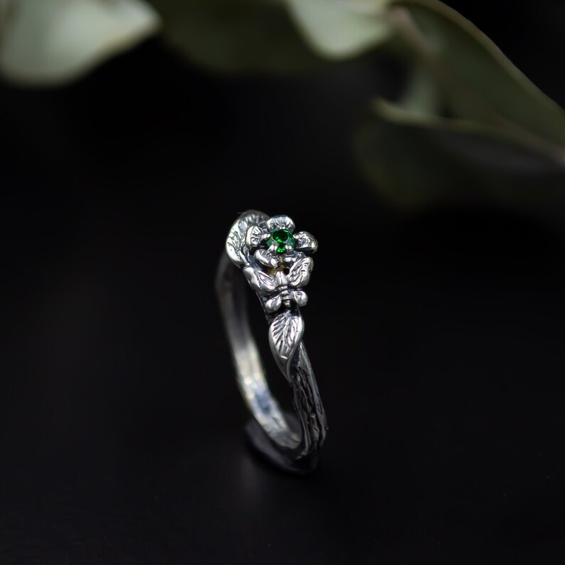 May include: A silver ring with a delicate floral design and a green gemstone. The ring is set on a black background.