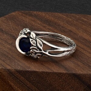 May include: A silver ring with a dark blue oval gemstone. The ring features a nature-inspired design with leaves and small accent stones. The band has a textured, bark-like appearance, and the ring is set on a wooden surface.