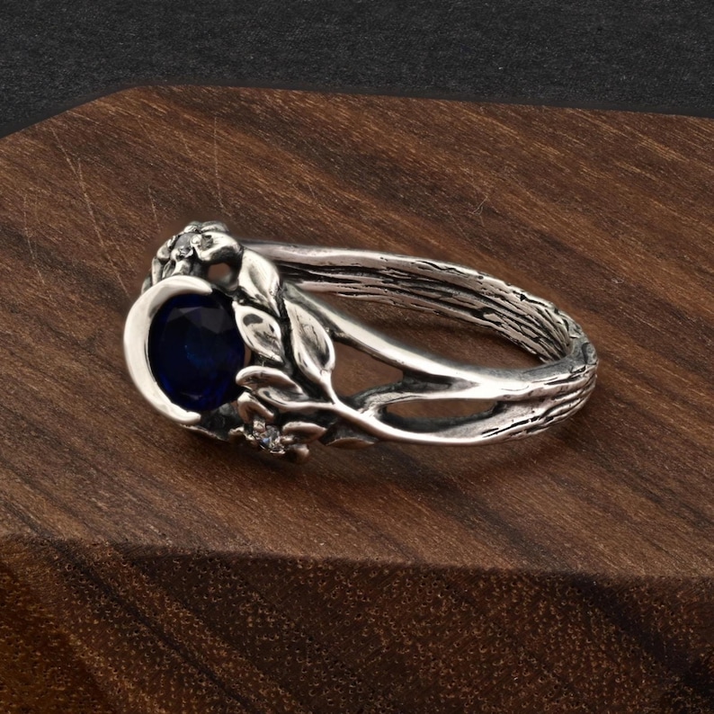 May include: A silver ring with a dark blue oval gemstone. The ring features a nature-inspired design with leaves and small accent stones. The band has a textured, bark-like appearance, and the ring is set on a wooden surface.